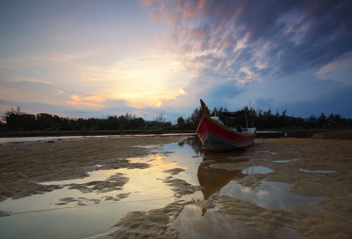 Free Serene beach scene with a boat reflected in water as the sun sets, creating a peaceful atmosphere. Stock Photo