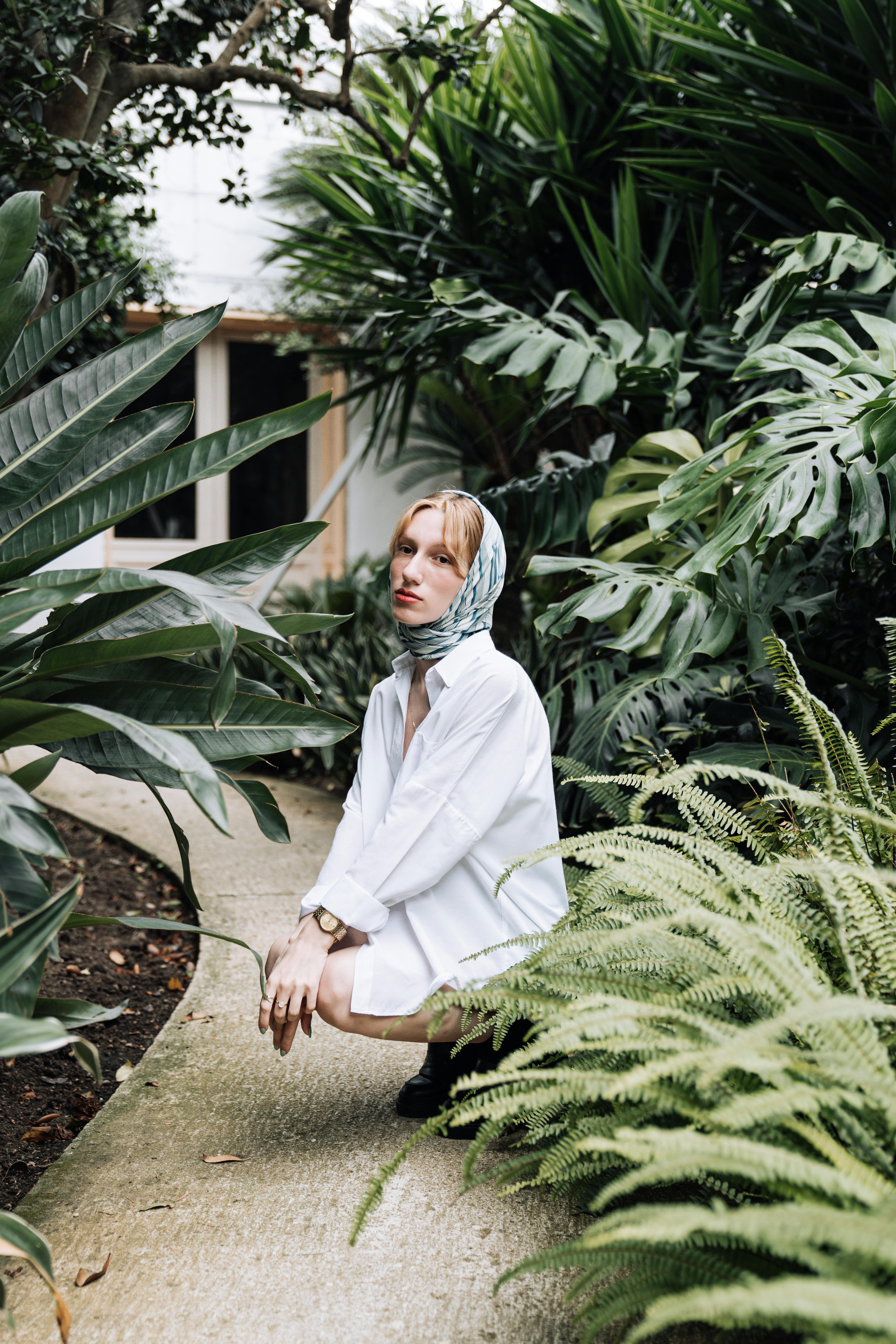 A young woman in a white blouse with a headscarf poses in a lush garden setting.
