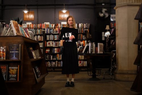Free Woman in bookstore holding a book surrounded by shelves of novels, creating a cozy and intellectual atmosphere. Stock Photo