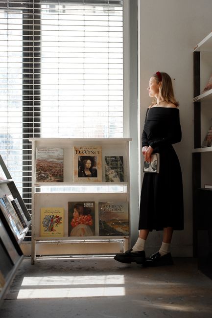 A young woman in a black dress stands in a sunlit bookstore, holding a book.