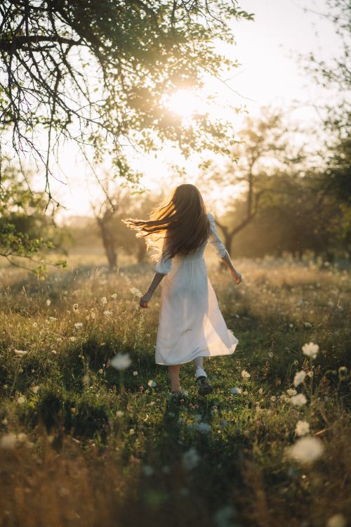Free A young woman in a white dress running joyfully through a sunlit meadow with trees. Stock Photo