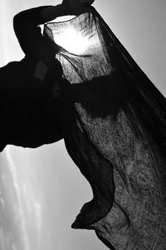 Dramatic silhouette of a woman holding fabric against the sun in monochrome.