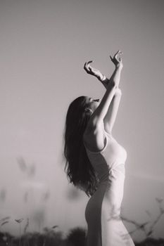 A young woman gracefully poses outdoors in a sleeveless dress, captured in monochrome.