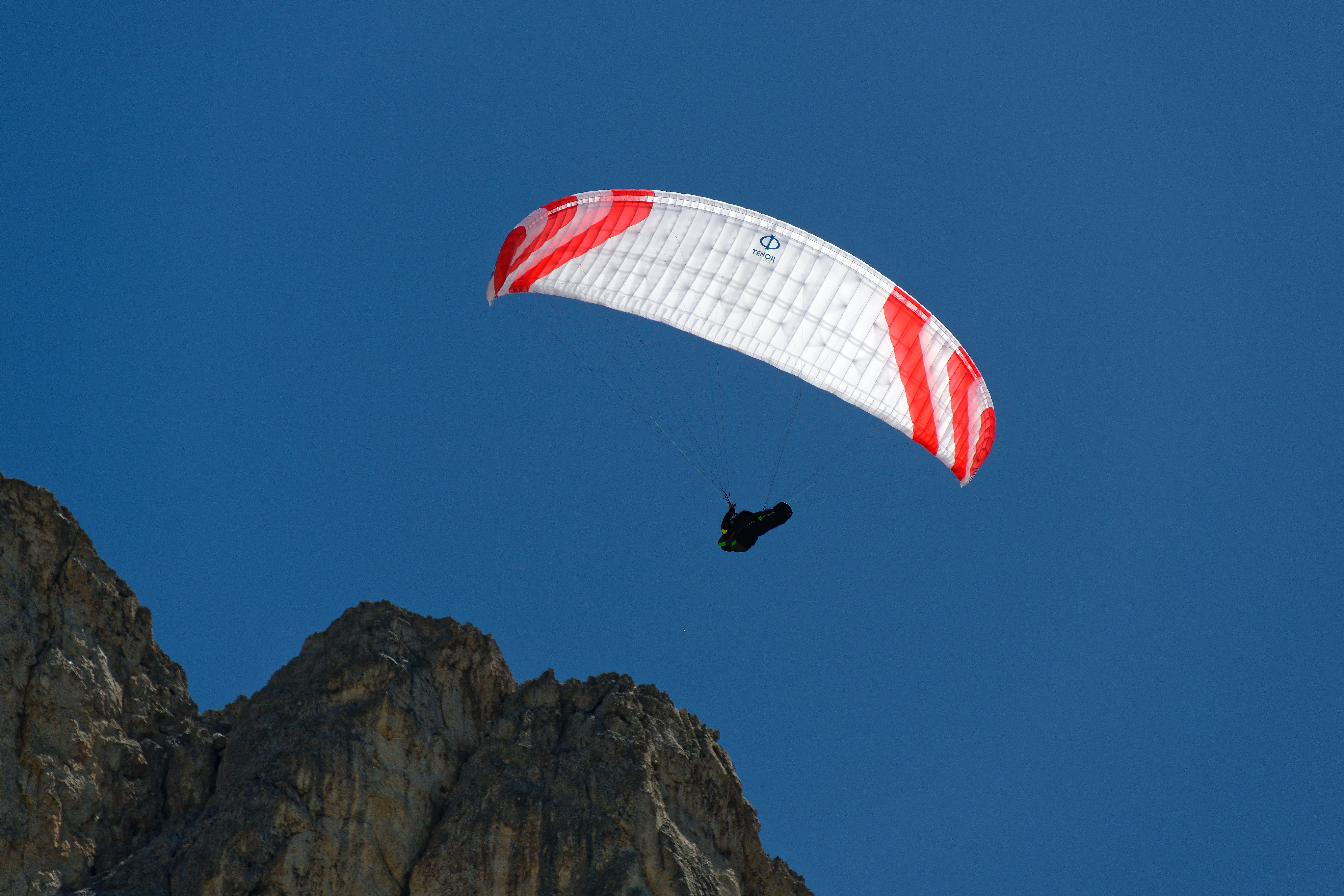 Paraglider flying over rocky mountains in Welschnofen, Italy, under a clear blue sky.