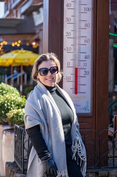 Fashionable woman in sunglasses standing by an outdoor thermometer on a sunny day.