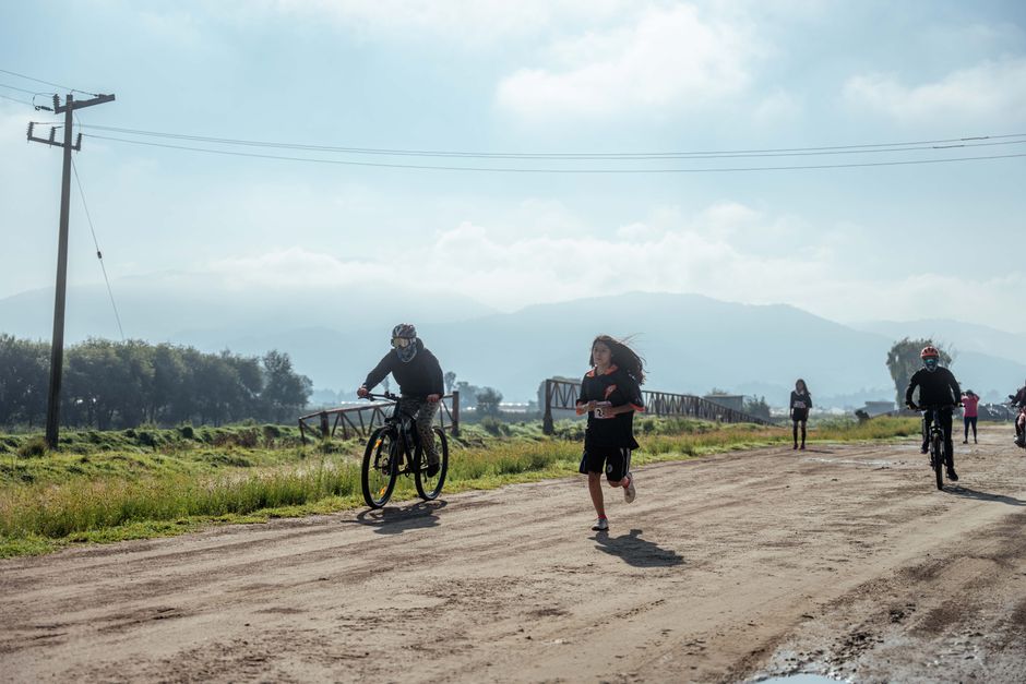 A group of people biking and running on a sunny day on a rural dirt road with mountainous backdrop.