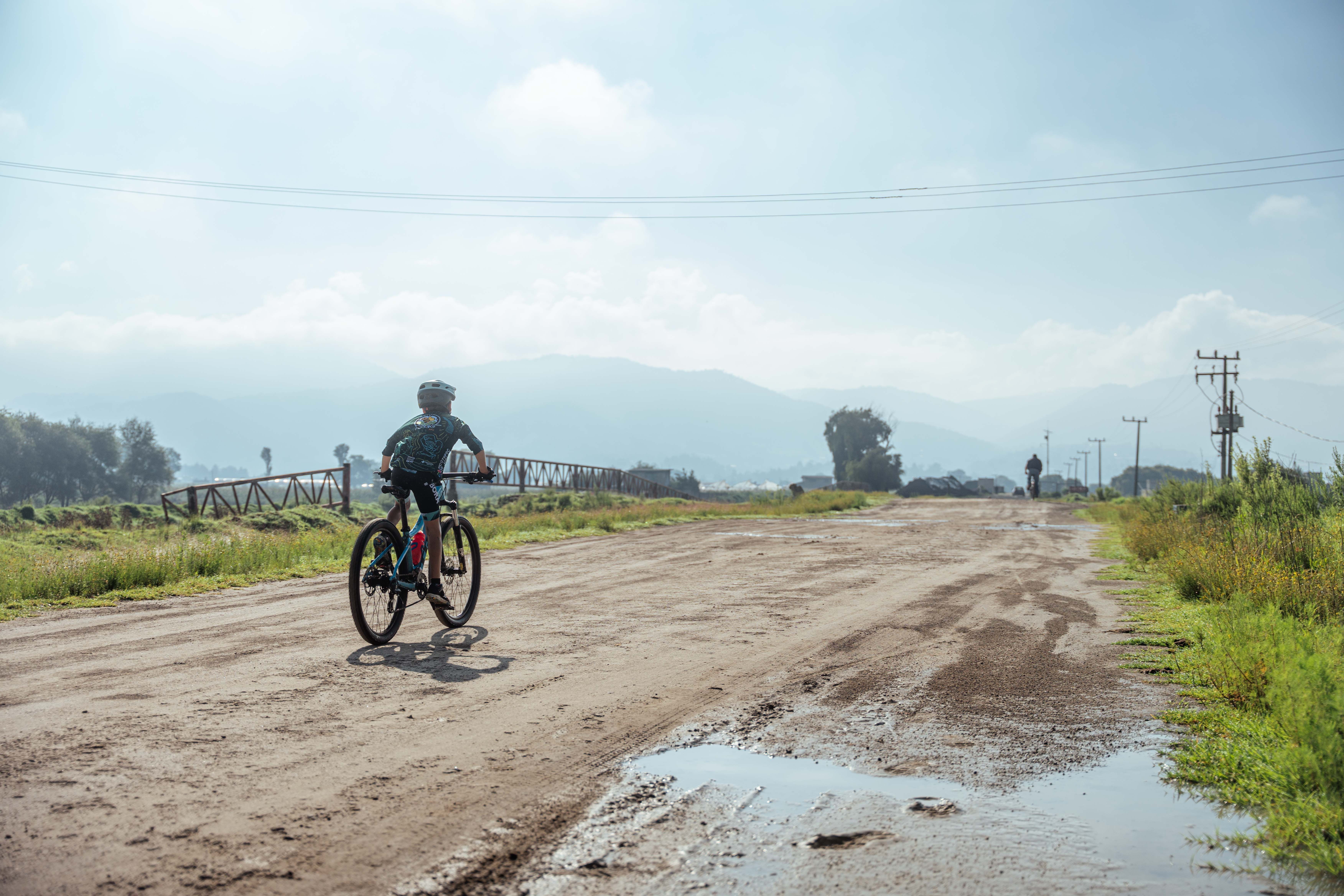 A child wearing a helmet rides a bicycle on a rural dirt road under a clear blue sky.