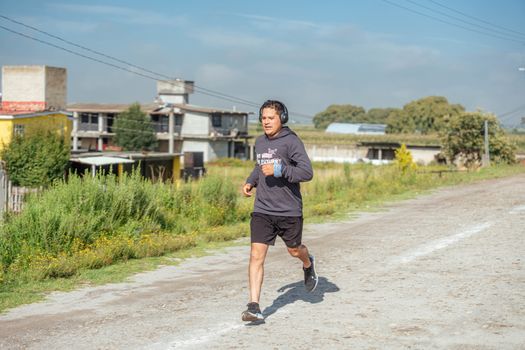 An adult man in a hoodie runs on a rural dirt road, listening to music with headphones.
