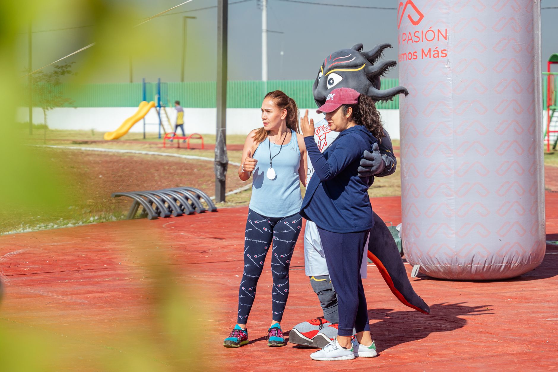 Women posing with a mascot at an outdoor sports event on a sunny day.