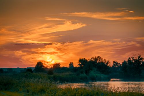 Free A peaceful countryside landscape featuring a vibrant sunset reflecting over a calm body of water. Stock Photo