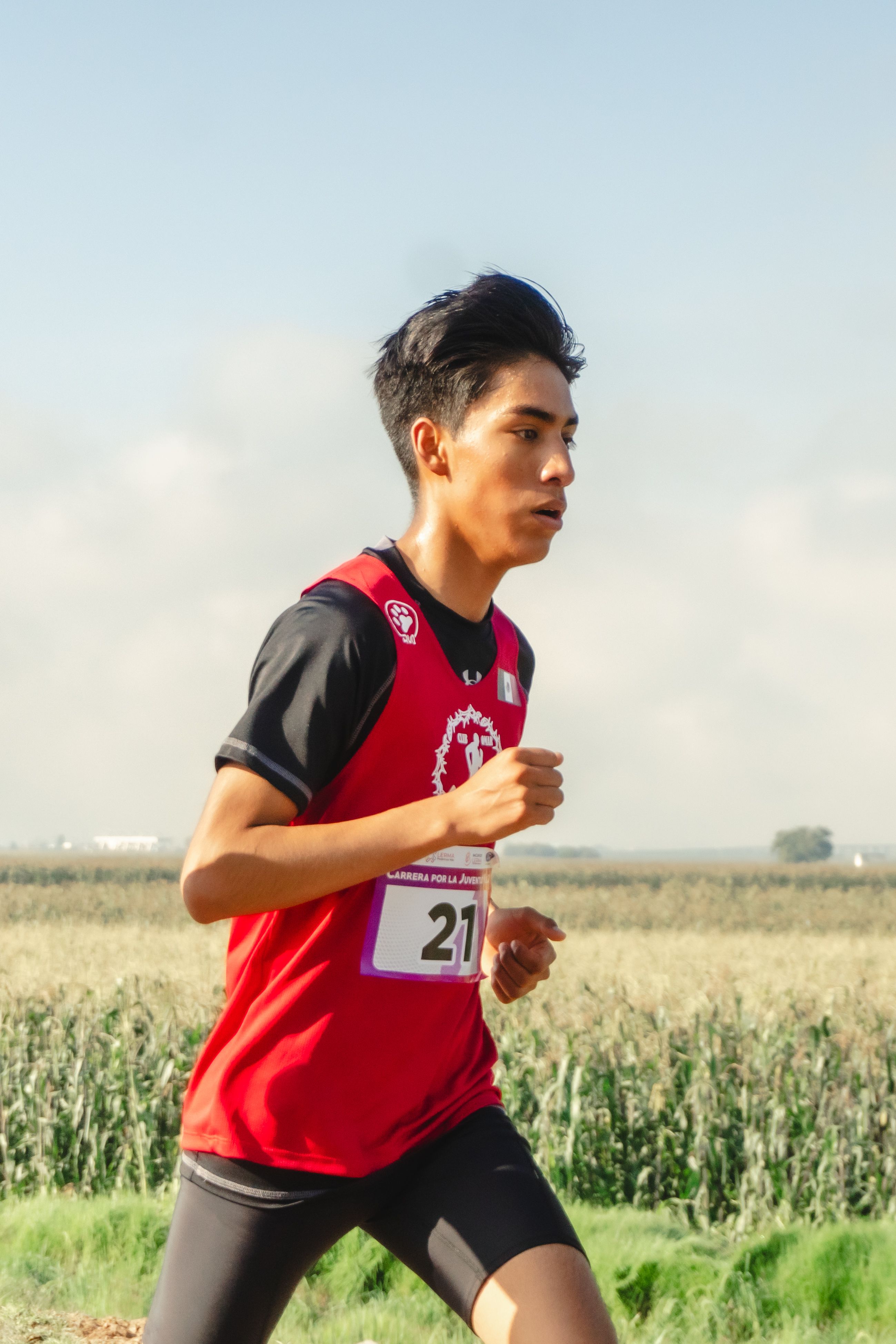 Hispanic male athlete running in a rural field during daylight, wearing sports gear.