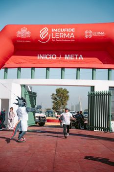 A man runs towards a finish line with a mascot cheering, during a sports event outdoors.