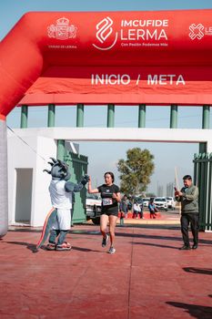 A woman runner celebrates finishing a race with a mascot at the finish line under a red arch.