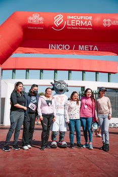 A group of women posing with a mascot under a sports event arch at the finish line, sunny day.