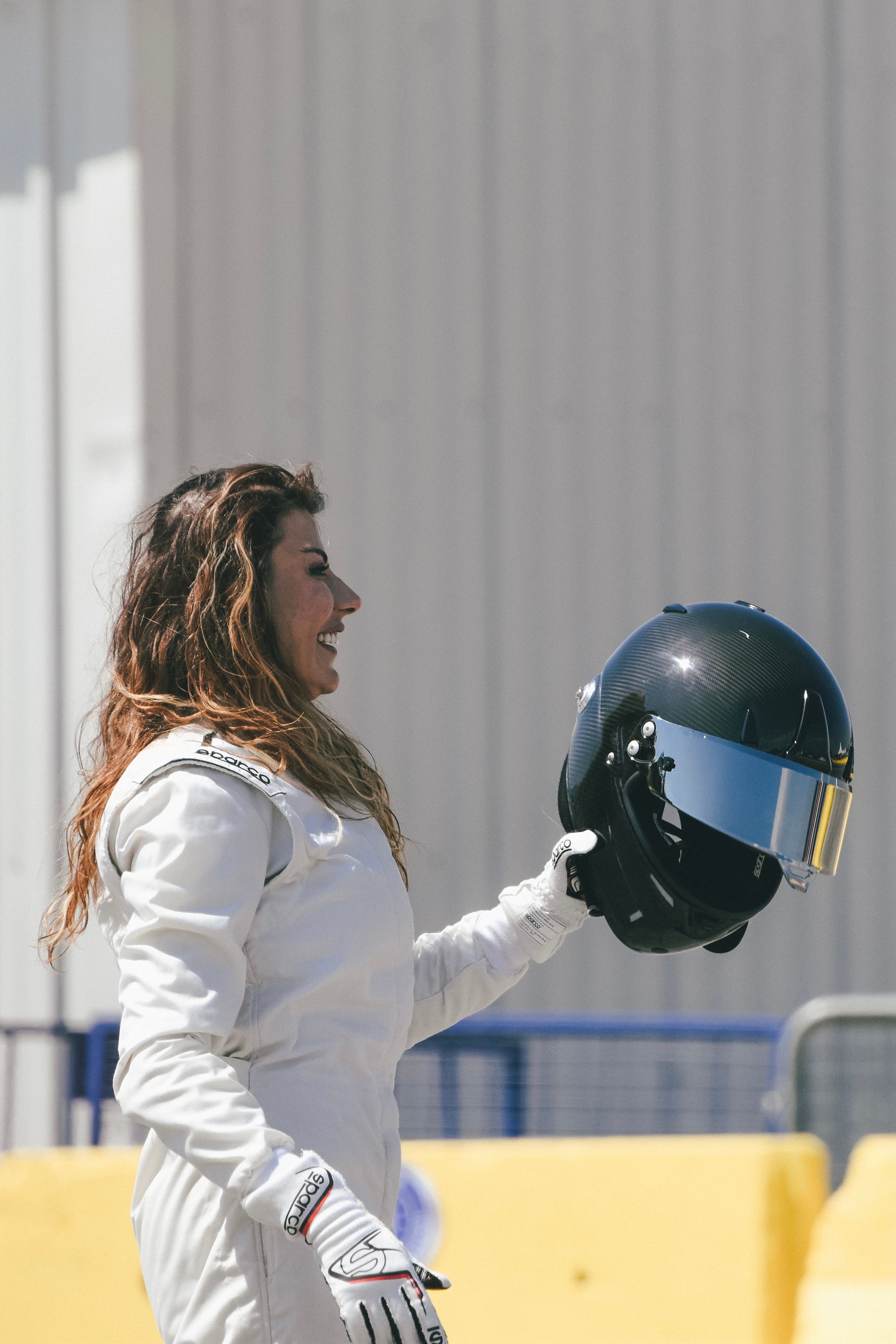 A female racecar driver with long brown hair holds a helmet while smiling outdoors.