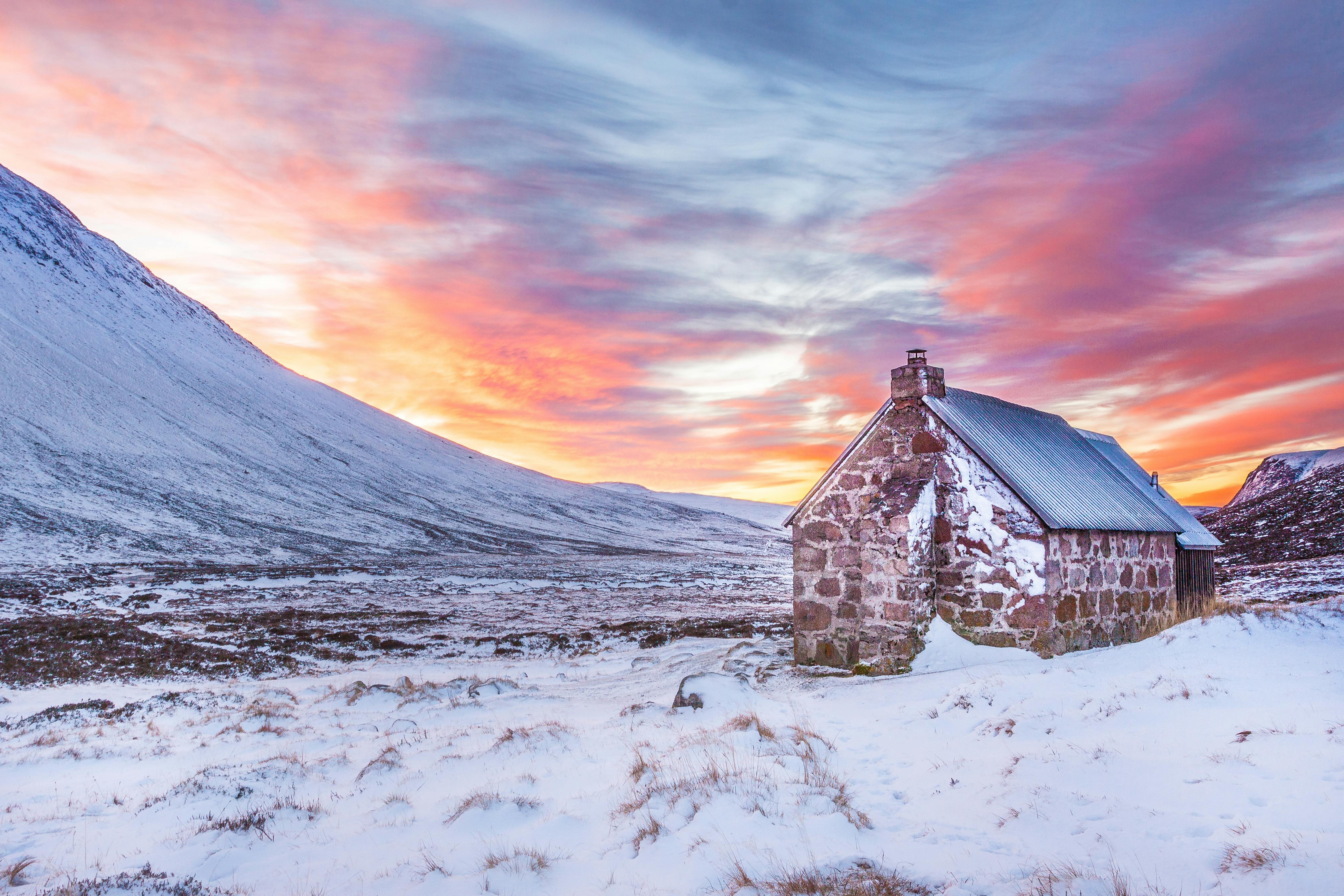 brown house surrounded by snow covered field near snow covered mountain under yellow blue and orange sunset