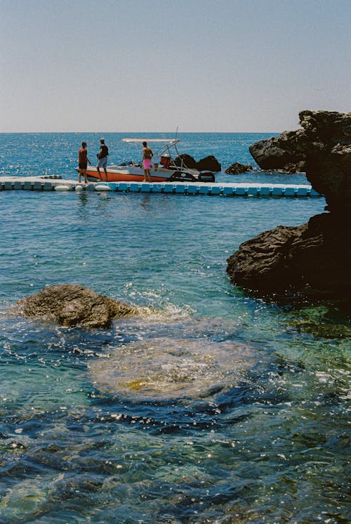 Free People enjoying leisure on a jetty with a boat by a rocky coastline during summer. Stock Photo