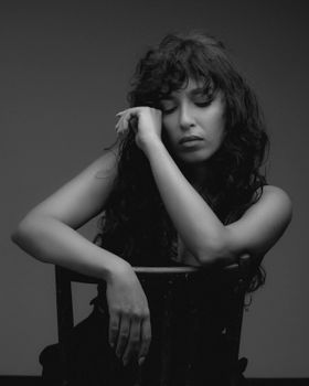 Moody black and white portrait of a woman with long wavy hair and closed eyes sitting on a chair.