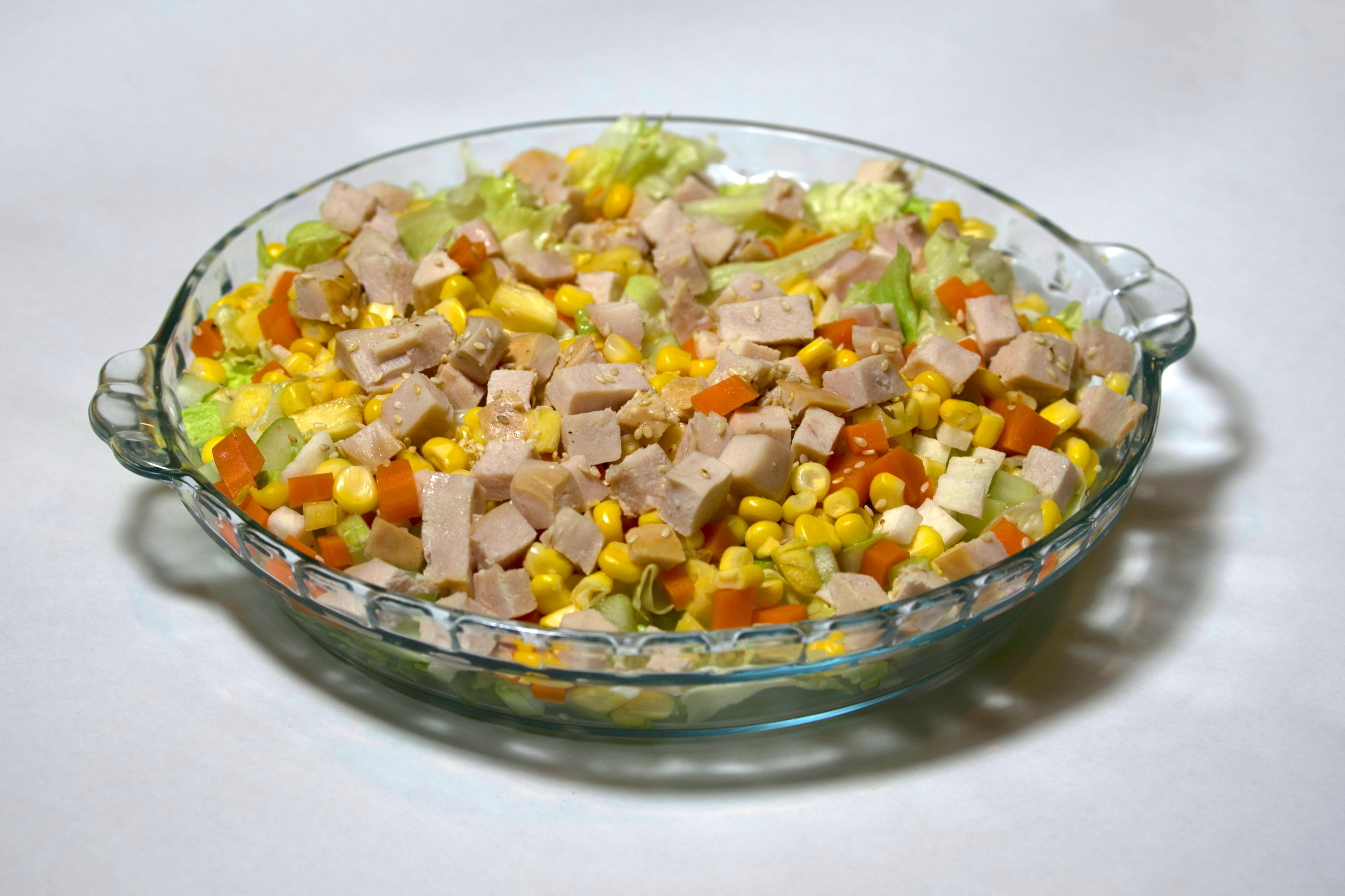 Vibrant mixed salad in a glass bowl featuring diced meat, corn, and vegetables on a white background.