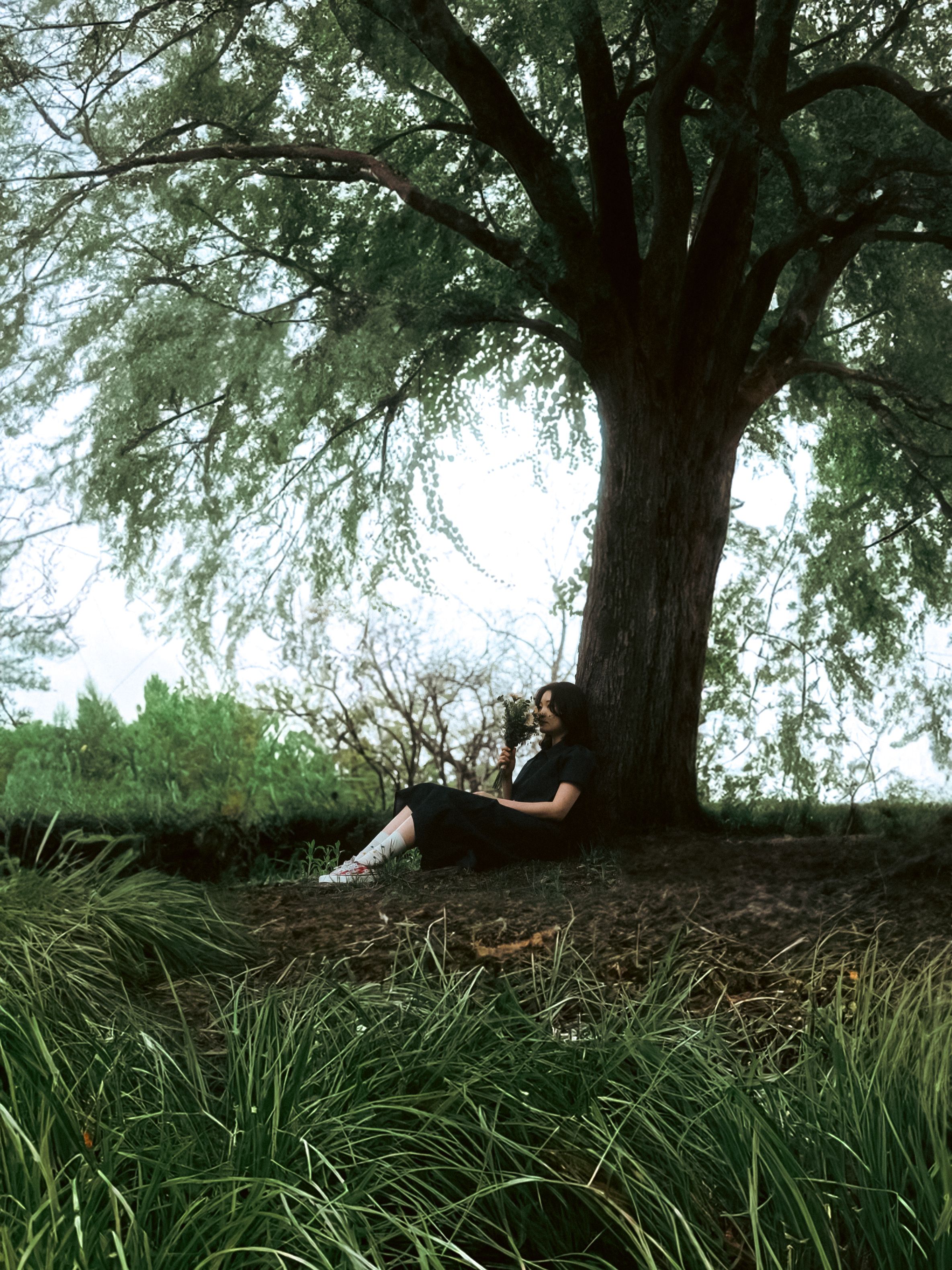 Adult woman sitting under a large tree in a meadow, enjoying a peaceful moment outdoors.