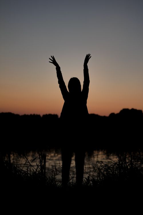 Free Silhouette of a woman with raised arms at sunrise by a serene lakeside. Stock Photo