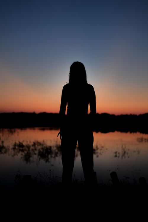 Free A woman stands silhouetted against a serene sunset by a lake, capturing peace and tranquility. Stock Photo