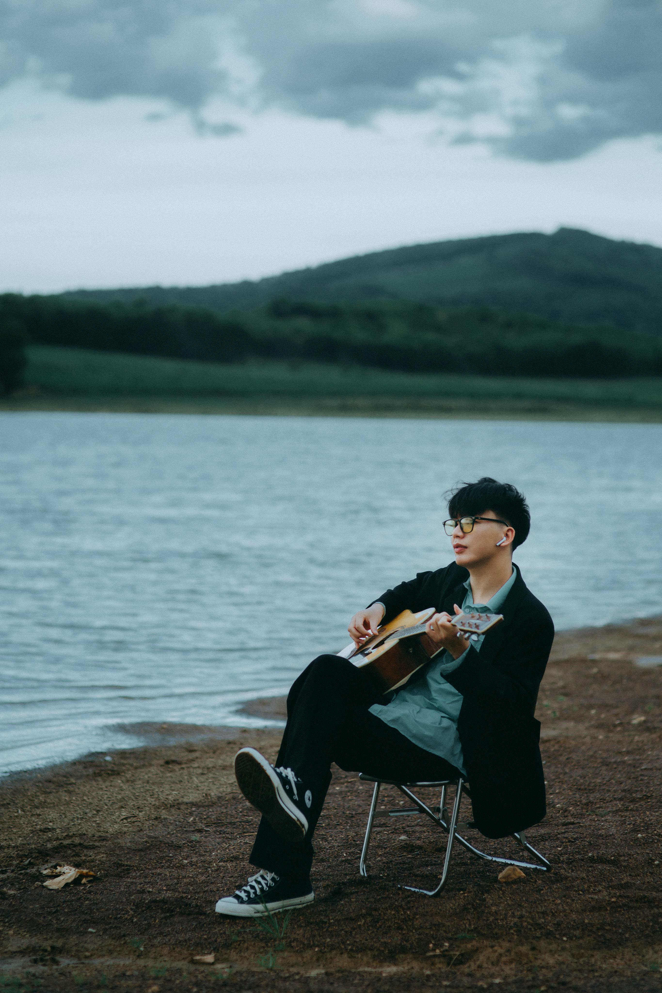 A young adult playing guitar on a chair by a serene lakeside, under a cloudy sky.