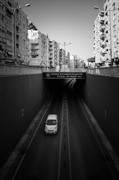 Black and white photo of a city tunnel with a car driving through and urban buildings above.