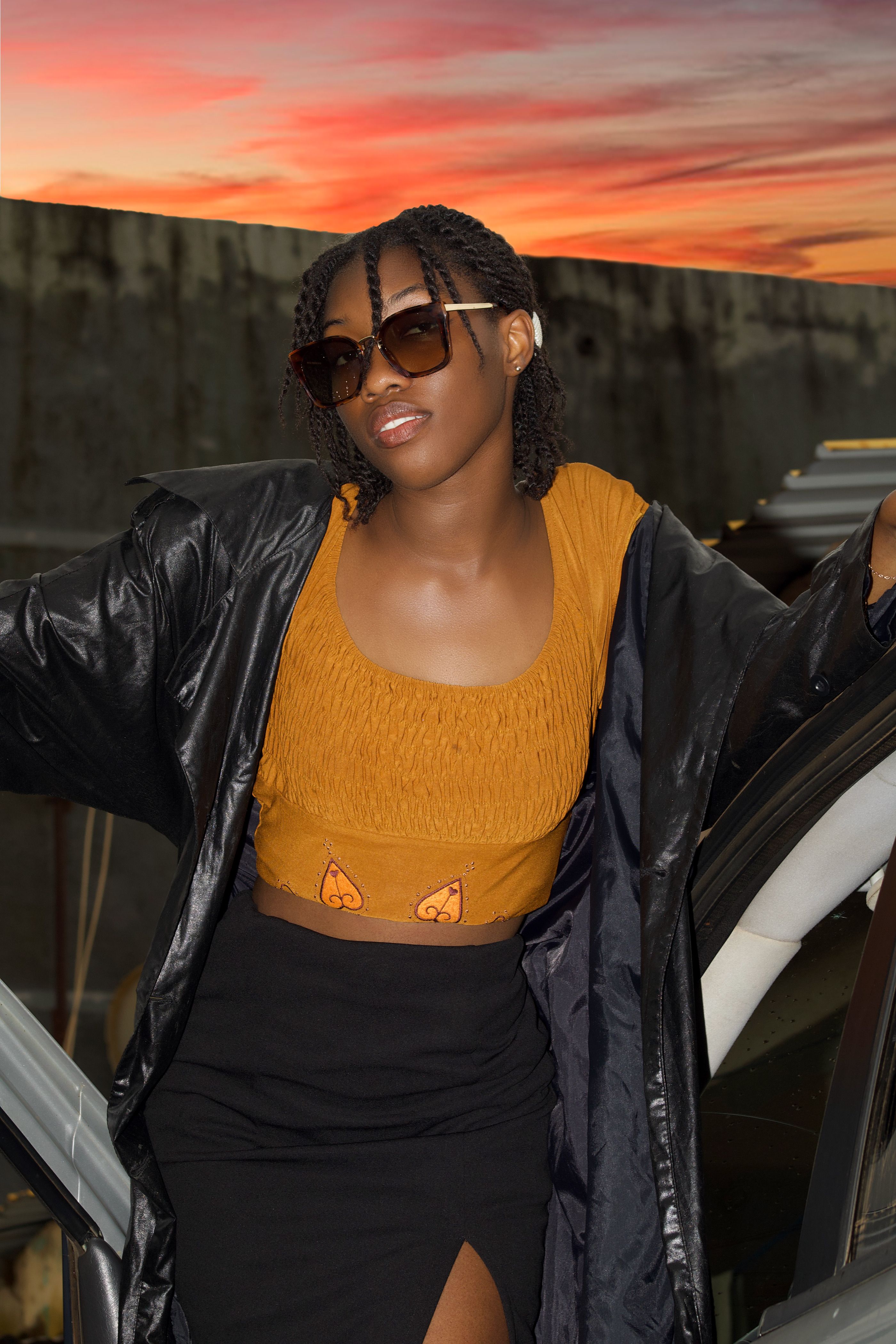 Mulher fashionista em casaco preto e blusa amarela posa ao pôr do sol, exudando confiança.