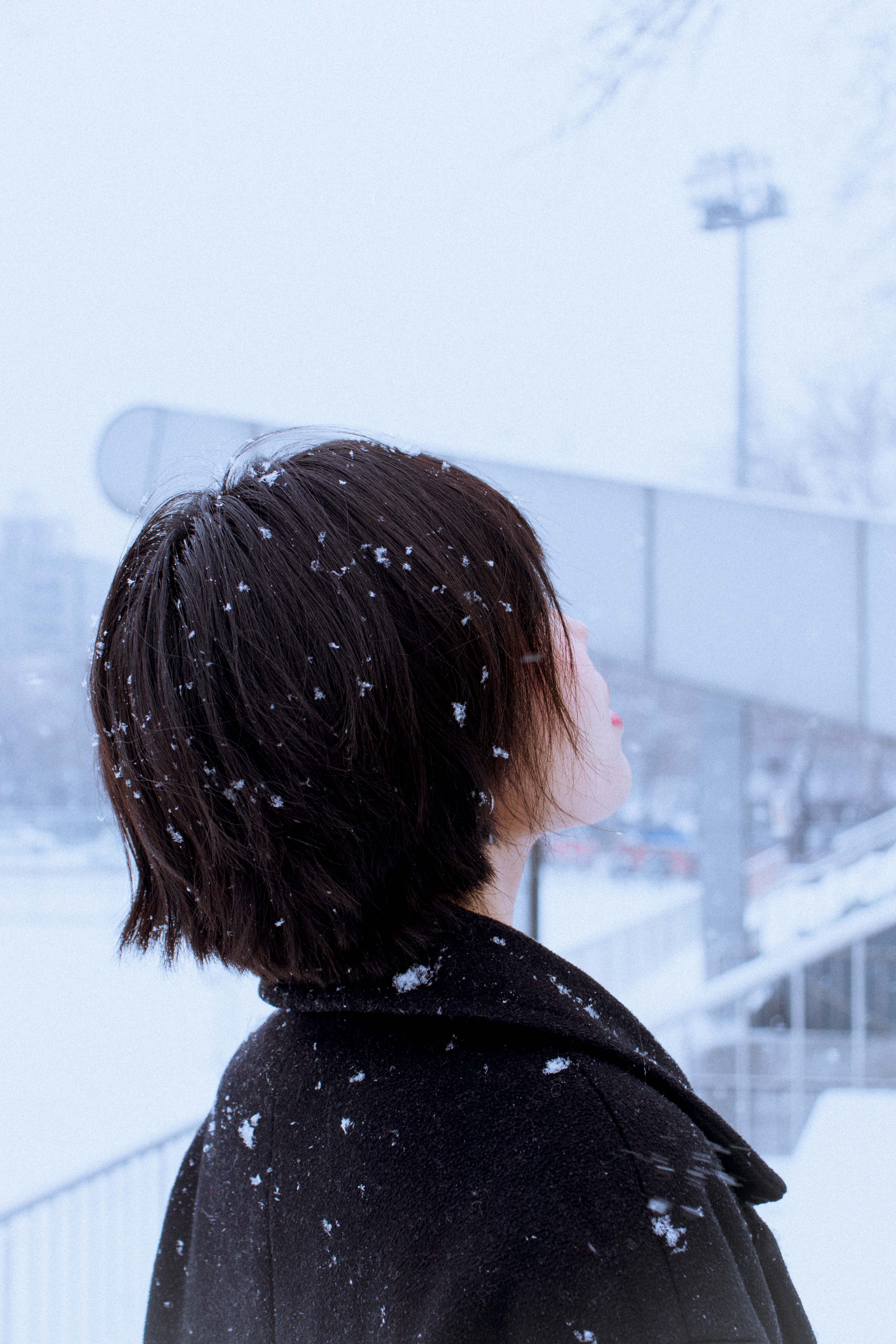 A woman stands in a snowy urban setting, gazing upwards as snowflakes fall around her.