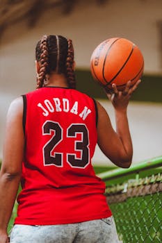 Woman in red sports jersey holding a basketball indoors, seen from behind.