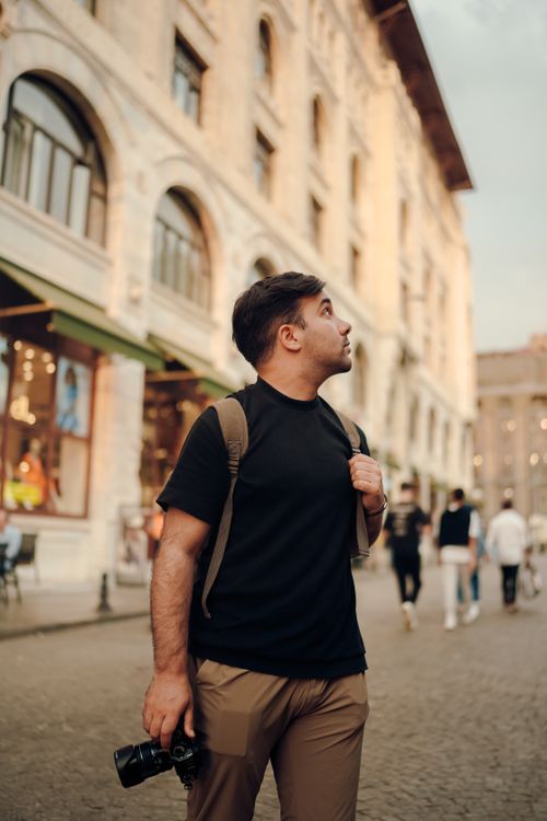 Free Man with camera walks through historic city streets, displaying urban architecture and travel vibes. Stock Photo