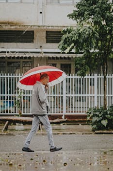 Adult man walks under a red umbrella along a rainy urban street, embodying a sense of urban resilience.