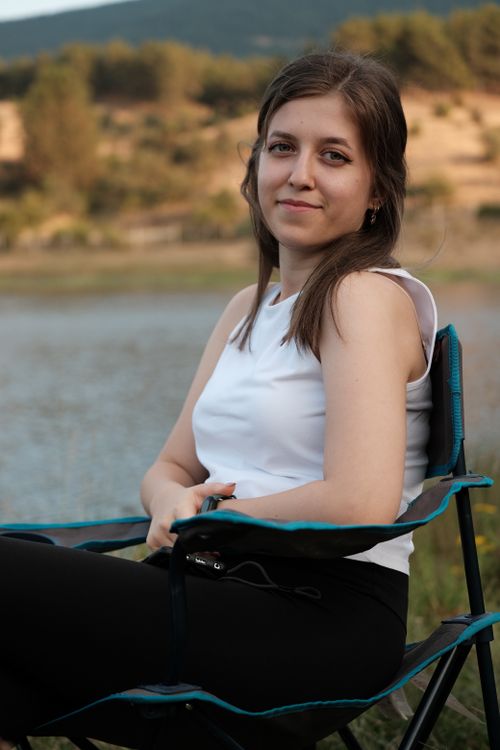 Free Young woman sitting on a folding chair near a peaceful lake, enjoying a summer day. Stock Photo
