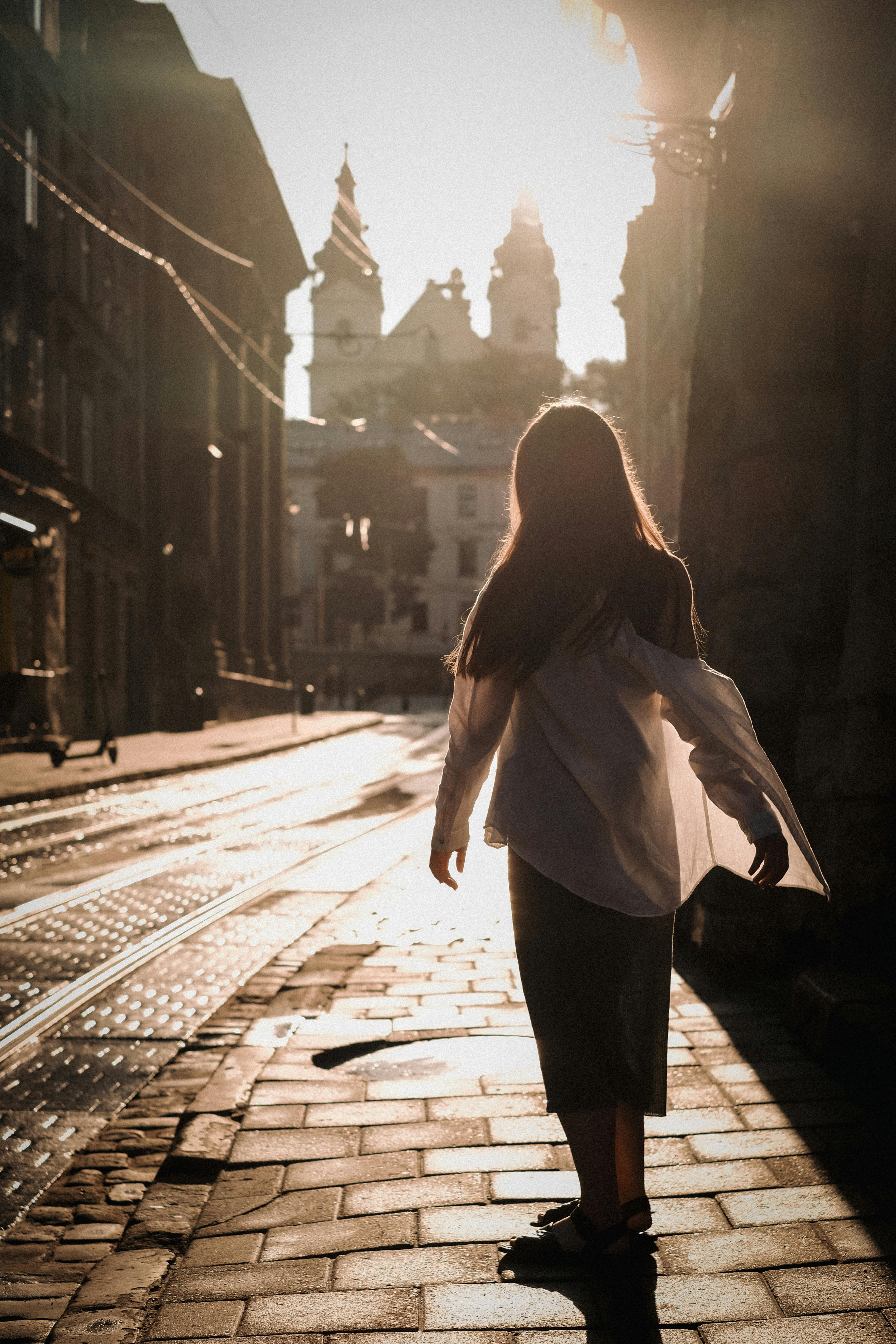 Silhouette of a young woman walking on a sunlit city street with historic architecture in the background.