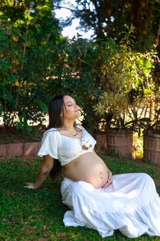 Pregnant woman in white dress sitting outdoors in a garden, enjoying the sunlight.