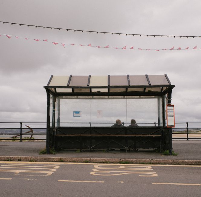 Two people sitting at an outdoor bus stop under cloudy skies, with road markings visible.