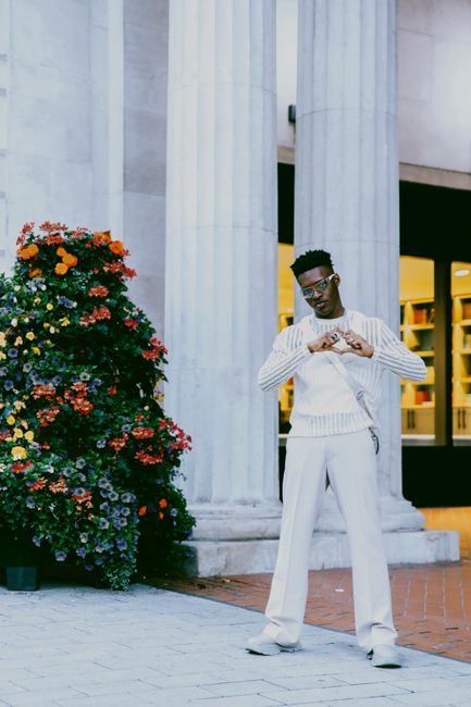 Fashionably dressed man poses by architectural columns with vibrant flowers.