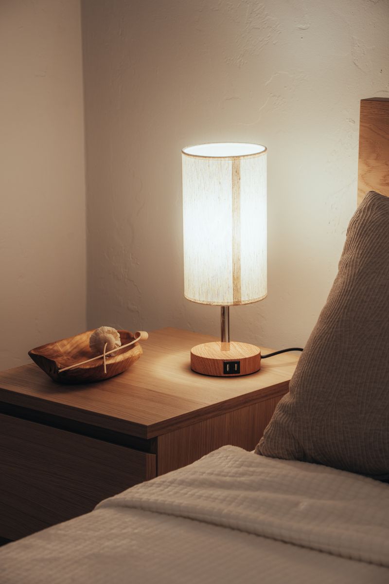 A lit lamp on a wooden nightstand beside a bed, photo by Osmany Mederos on Pexels