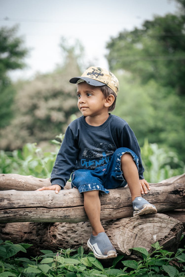 Boy In Cap Sitting On Tree Trunk In Forest