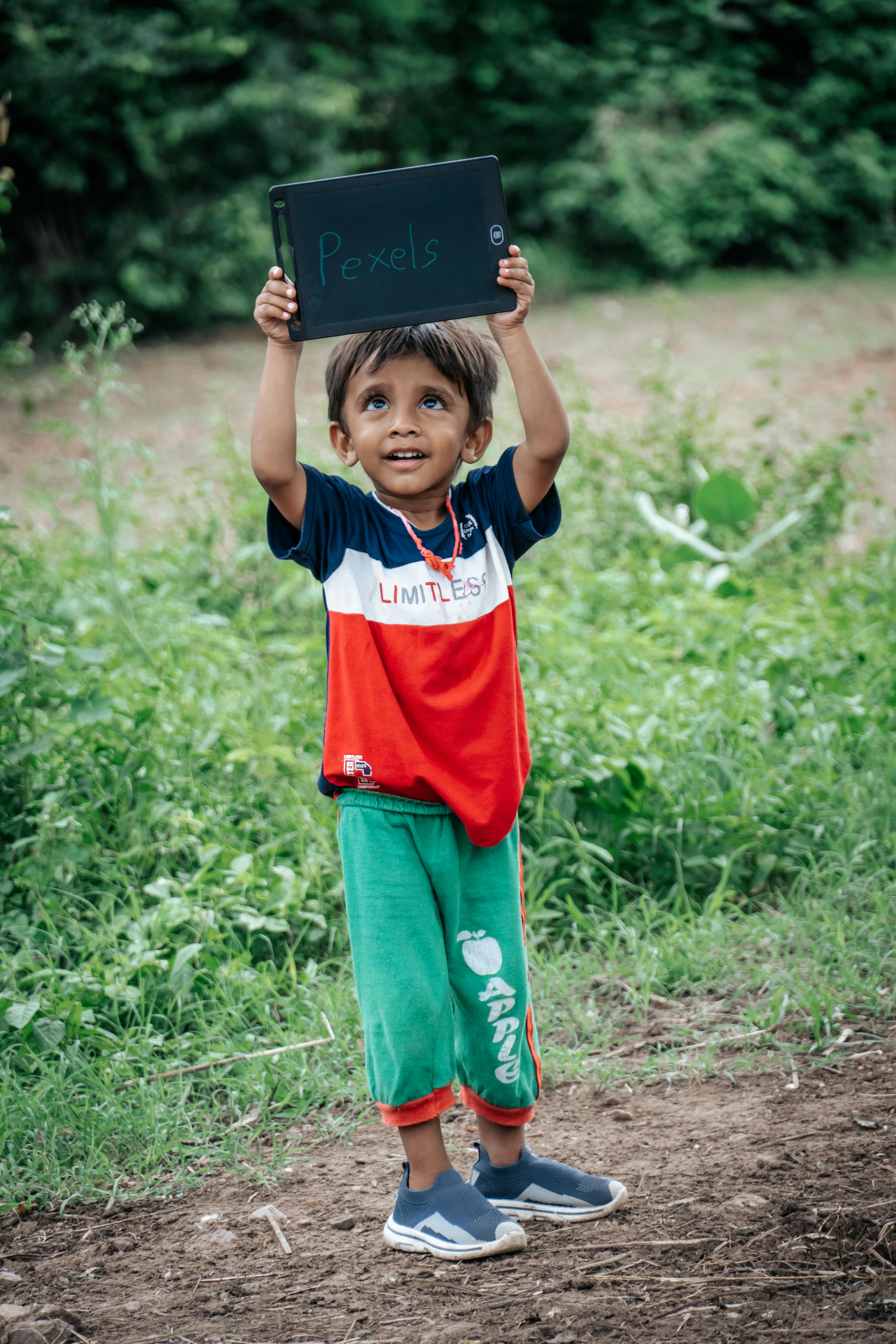 Young South Asian boy joyfully holding a tablet in a green outdoors setting.