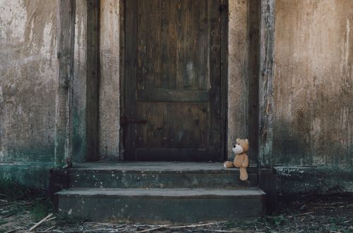 Free An abandoned teddy bear sits on steps in front of an old rustic wooden door. Stock Photo