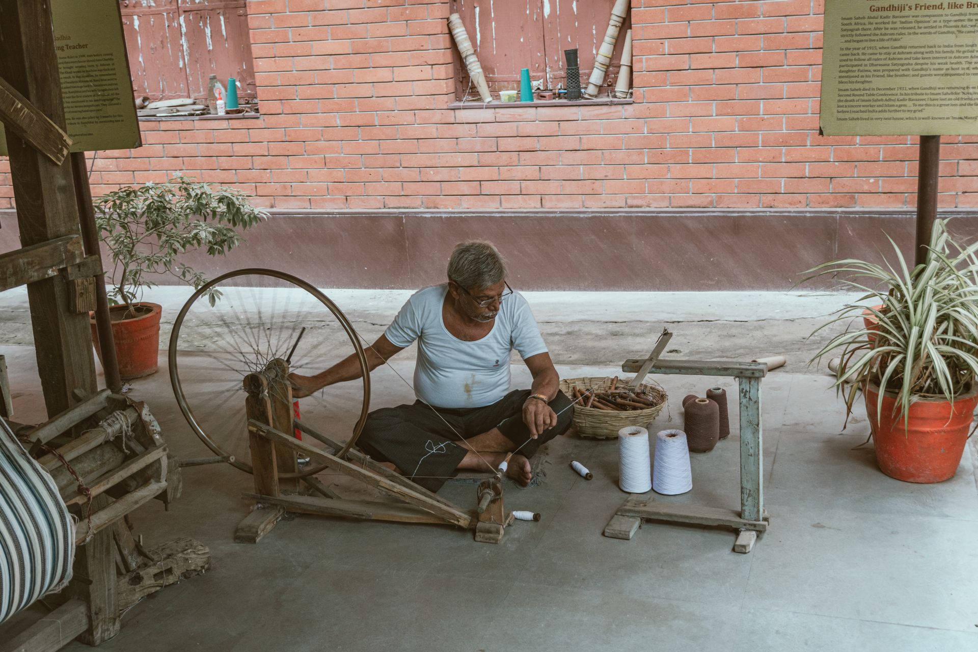 Elderly man sitting indoors, weaving thread on a traditional spinning wheel, surrounded by tools.