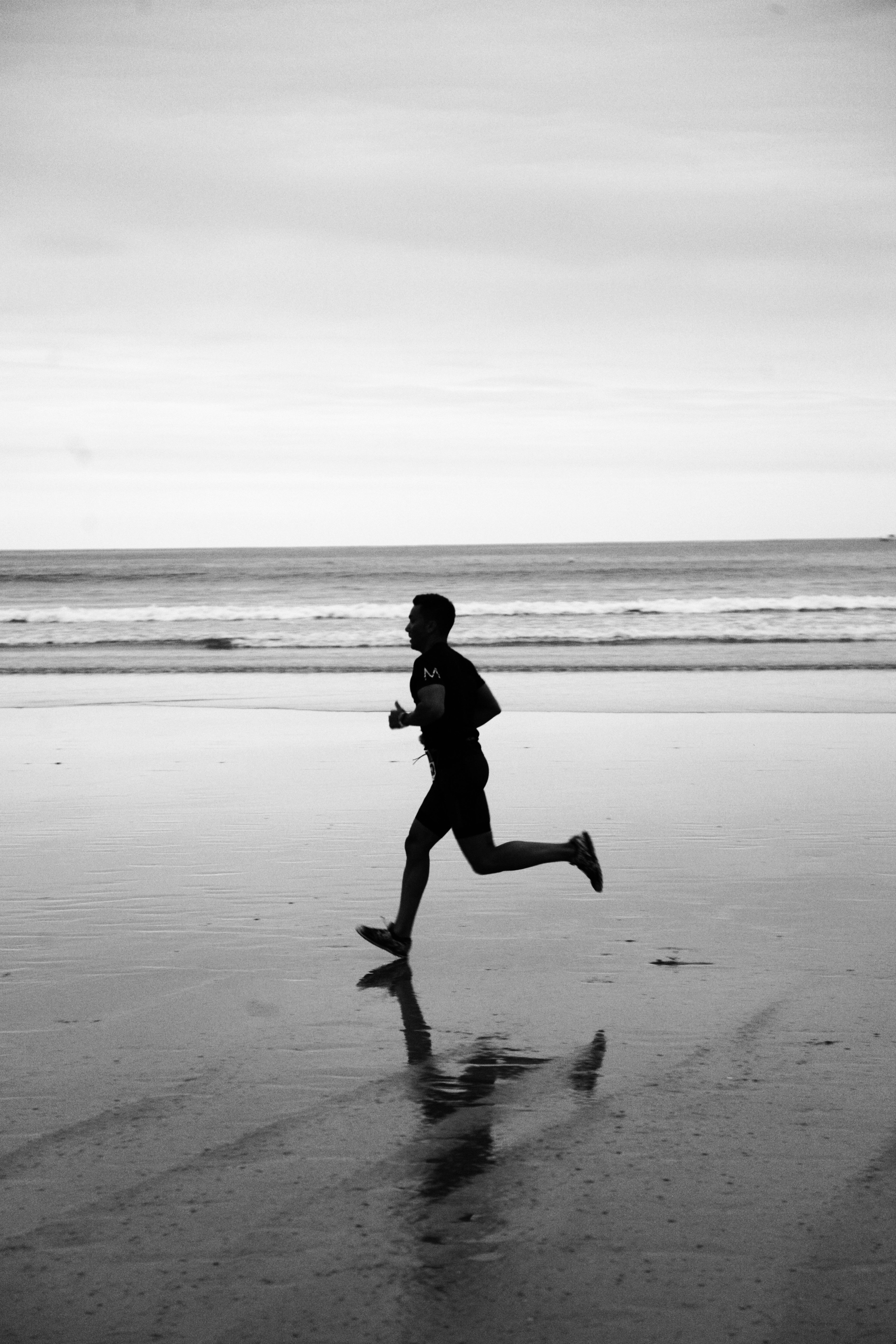 Black and white photo of a man jogging along the beach, reflecting health and leisure.