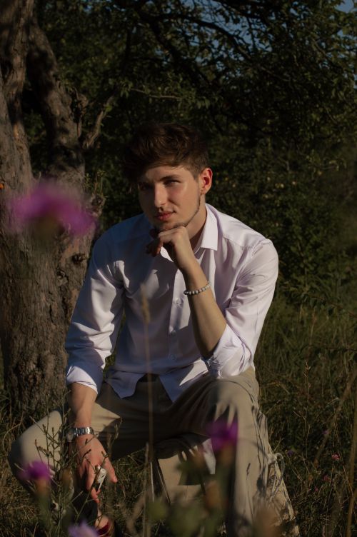 Free A young man seated outdoors in thought, wearing a white shirt amidst a natural green backdrop. Stock Photo