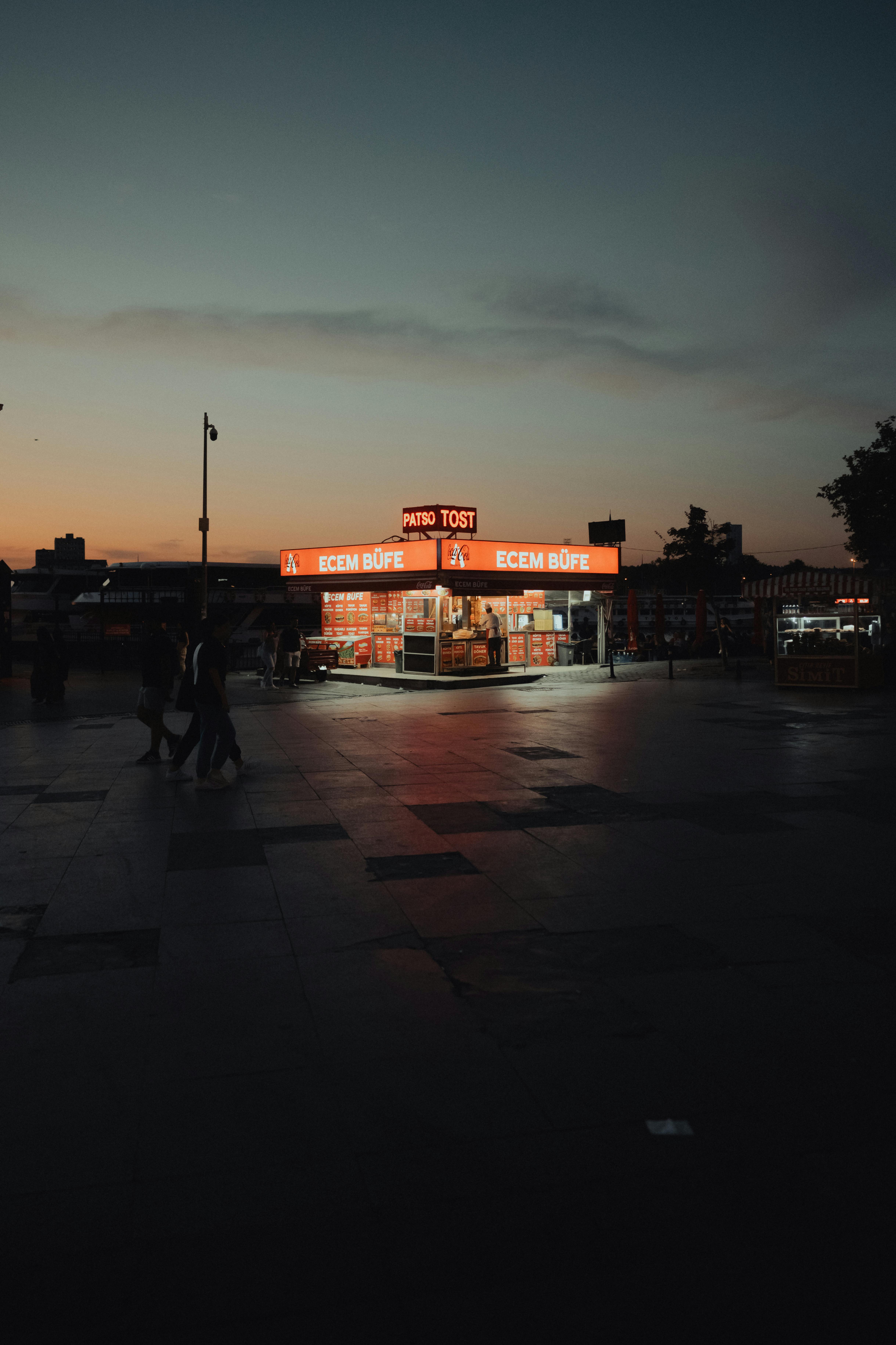 A brightly lit food kiosk in a city square during twilight creates a vibrant urban scene.