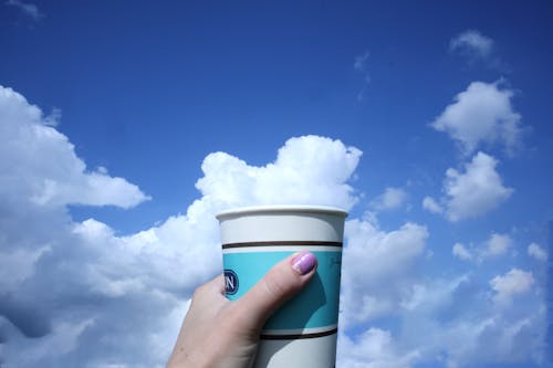 Woman Holding a Disposable Cup on the Background of a Blue Sky and White Clouds 