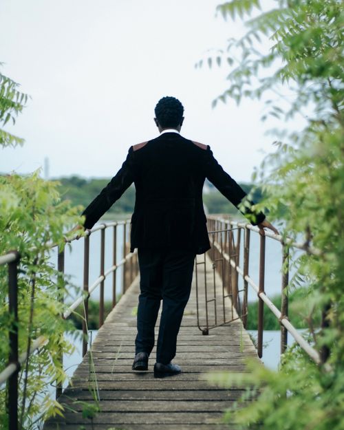 Free A man in formal attire walks across a wooden footbridge surrounded by lush greenery on a summer day. Stock Photo