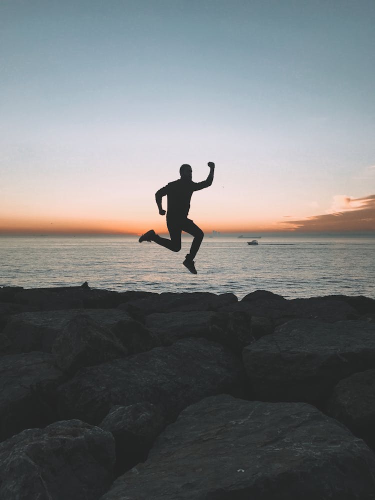 Silhouette Of Person Jumping Over Rocks On Seashore At Dusk