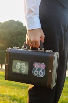 Close-up of a hand holding a retro suitcase with a Route 66 sticker in a sunlit outdoor setting.